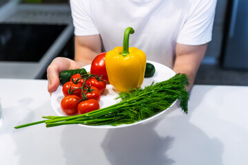the hands of a young man who holds a plate of fresh vegetables