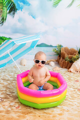 Happy little boy bathes in a bright inflatable pool on a sandy beach with palm trees by the sea