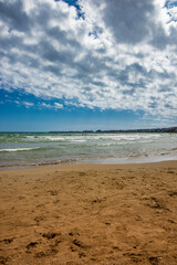 Nettuno, Lazio, Rome, Italy - The sea of the Roman coast, semi-deserted, at sunset. Cloudy blue sky in summer. The waves of the sea, slightly choppy. The beach, with sand and rocks.City in background