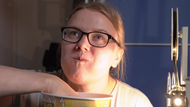 Young funny woman with glasses eats popcorn, greedily stuffing huge portions into mouth. A working vintage film projector is nearby. Concept of enjoyable home cinema experience.
