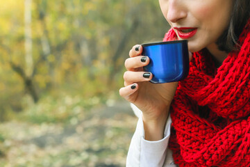 A woman in a white coat, crochet Red scarf and black hat holds a cup in her hands. Tea party in nature