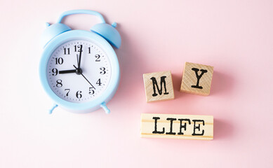 The text is my life on wooden blocks on a bed-pink background and a blue alarm clock.