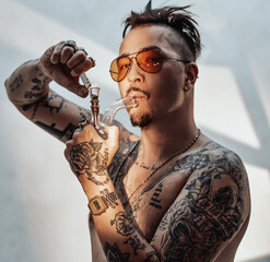 Shirtless gangster with tattooed body, smokes cannabis from bong