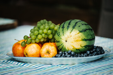 Plate of delicious fresh fruits like watermelon, apples, grapes and blueberries on a table a garden