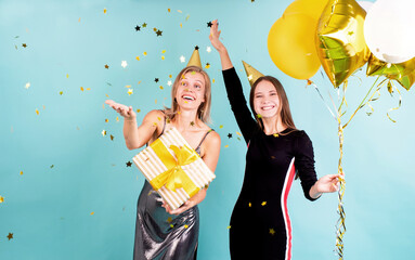 Two happy young women in birthday hats holding balloons celebrating birthday blowing confetti