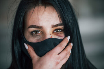 Portrait of beaten young woman in black protective mask with bruise under eye indoors in abandoned building