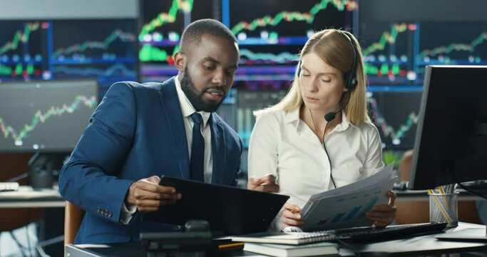 African American male trader asking gor advise in Caucasian woman broker in headset and with documents in hands. Mixed-races colleagues of stock market discussing rates. Multiethnic co-workers talking