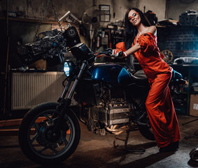 Fototapeta na wymiar Attractive sexual young woman in orange jumpsuit staying with a modern motorcycle in the car service