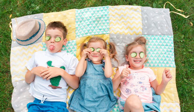 Kids Smile And Indulge Lying On A Patchwork Rug. Children Fooling Around With A Cucumber Mask. The Boy Shows His Tongue. Family Summer Camp Activity Ideas. Happy Kids Spend Their Summer Holidays.