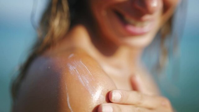 Close up of a happy smiling young woman is applying a sunscreen or sun tanning lotion on a shoulder to take care of her skin on a seaside beach during holidays vacation.
