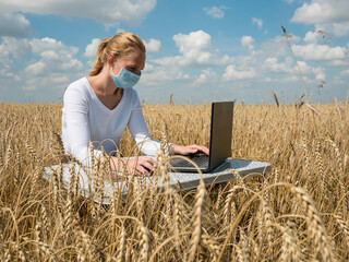 Naklejka na ściany i meble business woman at remote work. A young girl wearing a Covid-19 mask works for a laptop in a wheat field.