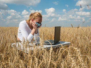 Naklejka na ściany i meble business woman at remote work. A young girl wearing a Covid-19 mask works for a laptop in a wheat field.