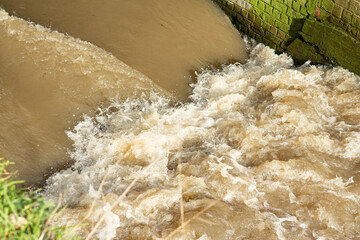 Close up on a little river. A difference in level causes swirling and foaming.The water is brownish, on one side a moss green brick wall, on the other a grassy bank.