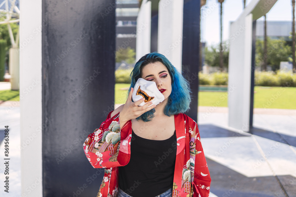 Poster closeup shot of a cool young lady with blue hair holding a mask in her hand posing in the street