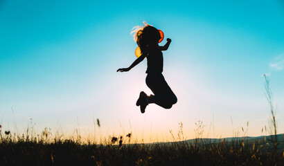 Blue yellow silhouette of a young woman jumping with balloons on a hill backlit by sunset light,...