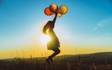 Silhouette of a child jumping with balloons on a hill backlit by sunset golden light, concept of...