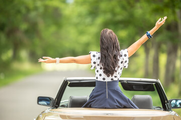 Woman sitting on top of cabriolet with arms open photographed from the back