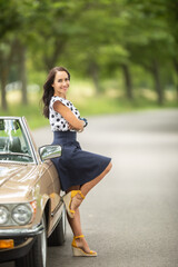 Beautiful woman in a skirt and high heels leans her back against a vintage convertible car parked...