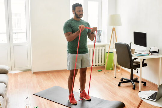 Tone up your body. Full length shot of young active man watching online video training on laptop, exercising with resistance band during morning workout at home