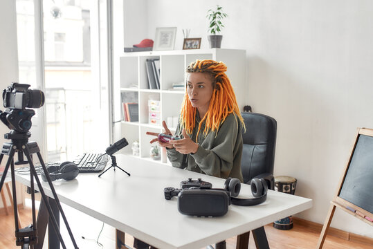 Female technology blogger with dreadlocks holding game controller joystick while recording video review of new gadgets using microphone at home