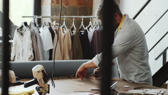 Caucasian male tailor cutting fabric with scissors and then folding piece while working on new custom clothes in dressmaking studio