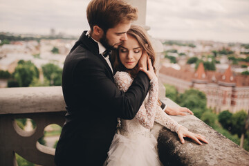 Happy newlywed. beautiful bride and stylish groom are hugging on the balcony of old gothic cathedral with panoramic city views