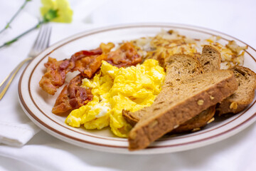 A view of a breakfast plate, featuring bacon, scrambled eggs, wheat toast, and shredded hash browns, in a restaurant or kitchen setting.