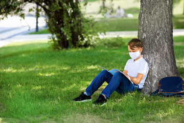 Naklejka na ściany i meble One boy wearing mask is sitting on the grass at park, reading book, doing home work after school during covid-19 pandemic time. New normal of coronavirus epidemic and education concept