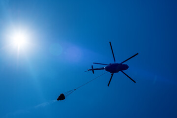 Fototapeta na wymiar Silhouette of a fire fighting helicopter with fire equipment on a blue sky with a radiant sun and natural lens flares