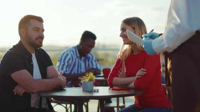 At sunlight happy young couple sitting at table outdoor in park at ordering dinner male waiter working food weekend guy selling retro retail shop order outside cafe slow motion