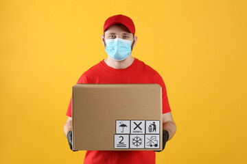 Fototapeta na wymiar Courier in mask holding cardboard box with different packaging symbols on yellow background. Parcel delivery