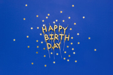 Festive candles in the form of the words Happy Birthday on blue background decorated with gold star confetti. Greeting card, top view