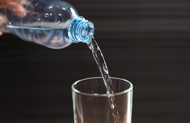 Pouring water from bottle into glass against dark background, closeup
