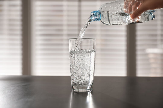 Person pouring water from bottle into glass on table against blurred background, closeup