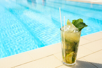 Glass of delicious mojito near swimming pool, space for text. Refreshing drink