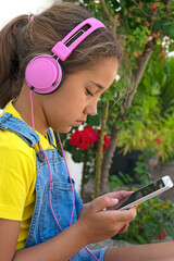 GIRL WITH FASHIONABLE HEADPHONES AND SMARTPHONE LISTENING MUSIC OUTDOOR IN THE GARDEN. URBAN SUMMER HOLIDAYS.