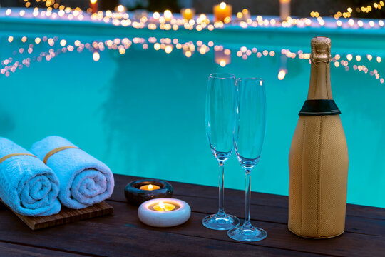 GOLDEN CHAMPAGNE BOTTLE AND TWO GLASSES WITH CANDLES AND TOWELS ON THE EDGE OF THE POOL. BACKGROUND WITH BLURRY LIGHTS
