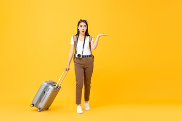 Full length portrait of smiling cute young Asian woman tourist with trolley bag opening hand to copy space aside isolated on yellow studio background