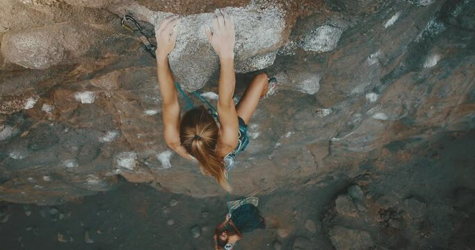 Beautiful young woman rock climbs mountain while friend belays her below, cinematic slow motion, fitness lifestyle