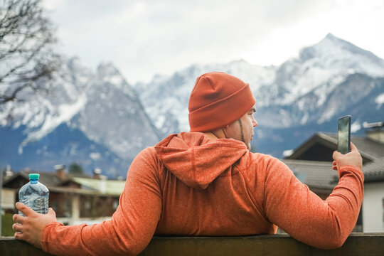 Outdoor Stylish man sitting on a bench, man in casual orange wear, stylish male wear hoodie and hat on Alps mountains background. european nature, relax, take it easy. garmisch-partenkirchen, Germany.