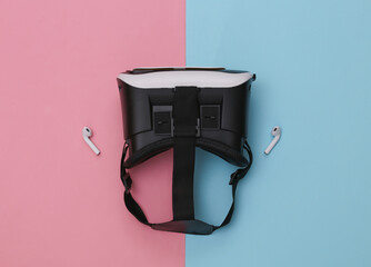 VR headset and wireless headphones on blue-pink pastel background. Modern gadgets. Top view