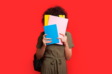 Caucasian student with curly hair covering her face with books and posing on a red studio wall...