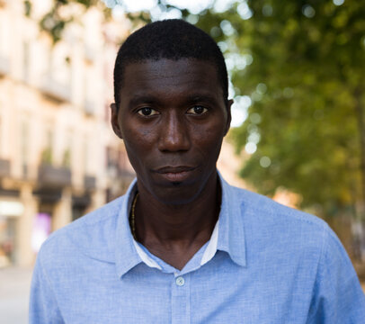 Headshot of young african american man wearing casual blue shirt looking confidently at camera against blurred background of summer city.