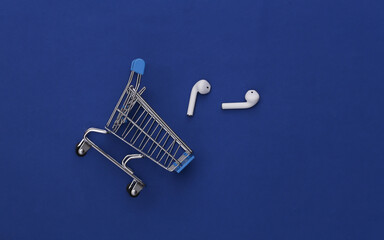 Shopping trolley and modern wireless earphones on classic blue background. Top view. Flat lay