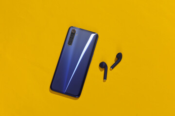 Smartphone and classic blue color true wireless bluetooth earbuds on bright yellow background. Top...