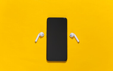 Smartphone and white true wireless bluetooth headphones or earbuds on bright yellow background. Top...