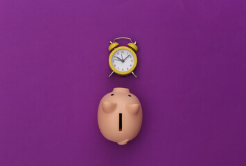 Time to invest. Piggy bank, alarm clock on a purple background. Top view. Flat lay