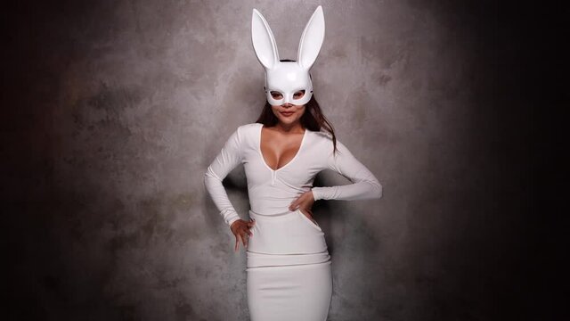 Portrait of beautiful flirting Asian woman in white dress and rabbit mask playfully posing for camera in the spotlight - video in slow motion
