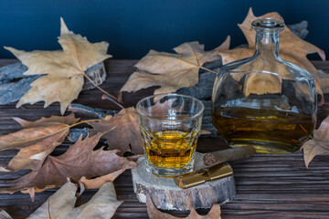 A glass of whiskey on a stand with a lighter and a cigar and a bottle of alcohol are on a table covered with leaves