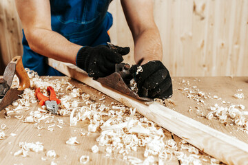 Close up of the man  in overalls treating a wooden bar with a black jack plane,  in the background...